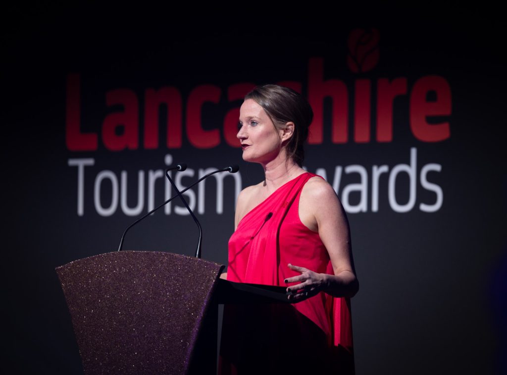The chief executive Rachel McQueen addressing the audience, from a lectern at the Lancashire Tourism Awards 2022 ceremony, A black tie event, she is wearing a red. on shouldered evening dress.