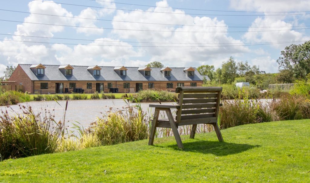 A view of Brickhouse Farm Holiday Cottages. A row of attractive red brick, wooden clad cottages overlooking a lake, trees and gardens with bench seating.
