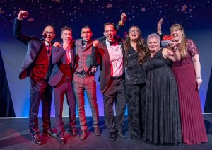 A group of six jubilant men and women from Brickhouse Farm Cottages, celebrate their win on stage. Joined by guest presenter, Steve Royle, punching the air.