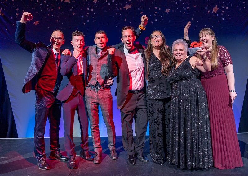 A jubilant group photograph of men and women from Brickhouse Farm Cottages celebrating their win on stage. Arms raised. The guest presenter, Steve Royle, joins them in punching the air. 