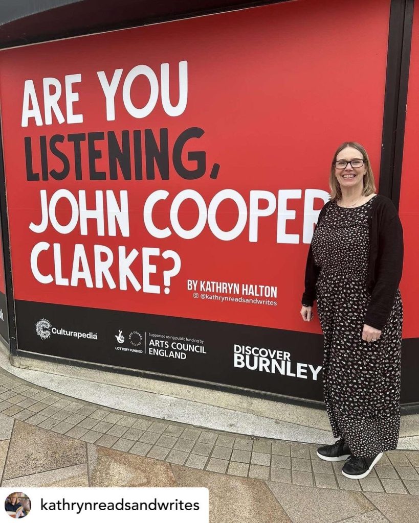 A shop front window covered with the large text 'Are You Listening John Cooper Clarke?" with Kathryn Halton standing to the side. The background of the sign is bright red with the words written in white and black. 