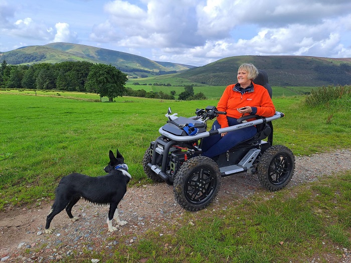 An all-terrain wheelchair with lady occupant is stationary, on a track in the Forest of Bowland, Lancashire. Trees and hills as far as can be seen, with blue skies and a smattering of clouds. The wheelchair user is accompanied by a dog which is on the path next to her.